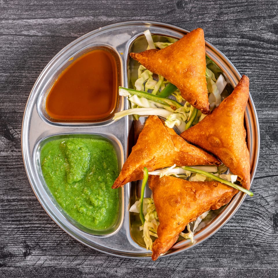 Four golden-brown samosas with shredded cabbage, green chutney, and tamarind sauce on a metal plate.